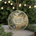 A round ornament with a painting of a cliff and a bird, surrounded by greenery, is displayed on a surface with Christmas lights in the background.