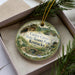 A decorative ornament in a box, featuring a scenic landscape with a bridge and trees, with the text "Carter Caves State Resort Park, Kentucky" printed on it.