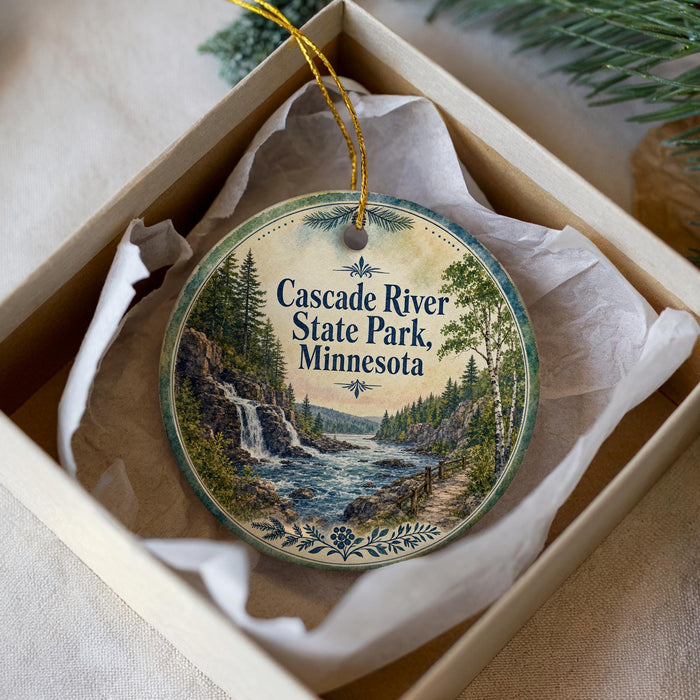 A round ornament with a scenic image of the Cascade River State Park in Minnesota, displayed in a box with a white tissue paper lining.