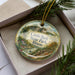 A decorative ornament in a box, featuring a scenic landscape with a castle, trees, and a river.