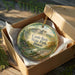 A decorative ornament in a box, featuring a scenic landscape with a castle, trees, and a river.