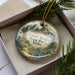 A decorative ornament in a box, featuring a scenic landscape with mountains and pine trees.
