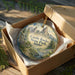 A decorative ornament in a wooden box, featuring a scenic landscape with mountains and a river.