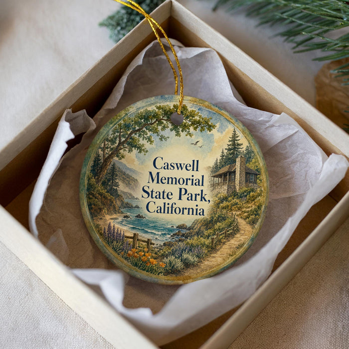 A decorative ornament in a white box, featuring a scenic landscape with a house, trees, and a path leading to a body of water.
