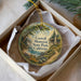 A decorative ornament in a white box, featuring a scenic landscape with a house, trees, and a path leading to a body of water.
