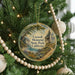 A round ornament hanging on a Christmas tree, featuring a scenic landscape with a river, mountains, and a cabin.