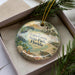 A decorative ornament with a beach scene and various sea creatures, including a bird, is displayed in a box with pine branches.