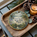 A decorative ornament with a scenic landscape of Chain O Lakes State Park, Illinois, featuring a bird and pine cones, placed on a wooden tray alongside a lit candle.