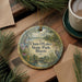 A decorative ornament with a scenic landscape of Chain O Lakes State Park, Illinois, featuring a bird and flowers, is displayed on a wooden surface alongside a cup of coffee and some pine branches.