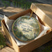 A decorative ornament in a cardboard box, featuring a scenic landscape with a lake, trees, and birds.