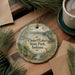 A decorative ornament featuring a scenic landscape with a lake, trees, and wildlife, including a deer, is displayed on a wooden surface alongside a cup of coffee and some papers.