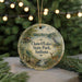 A round ornament with a scenic landscape featuring a lake, a cabin, and wildlife, including a deer, is displayed in front of a blurred background with Christmas lights.