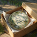 A decorative ornament in a wooden box, featuring a scenic landscape with a lake, trees, and wildlife.