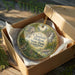 A decorative ornament in a cardboard box, featuring a scenic landscape with a bird and a waterfall.