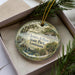 A decorative ornament in a box, featuring a scenic landscape with a body of water, trees, and a bird.