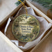A circular ornament with a painting of a pond and a bird, surrounded by greenery, placed in a box with a white tissue paper lining.