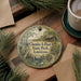A round ornament with a painting of a bird and a house, surrounded by pine branches and a cup of coffee on a wooden surface.