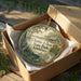 A circular ornament with a painting of a park scene, placed in a cardboard box with a string hanging from it.