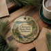 A decorative ornament featuring a scenic landscape of Chewala State Park, Alabama, with pine trees and a waterfall. The ornament is hanging on a wooden surface, accompanied by a cup of coffee and some papers.