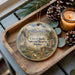 A decorative ornament with a scenic landscape of mountains and a wagon, placed on a wooden tray alongside pine cones and a lit candle.