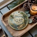 A decorative ornament with a lighthouse and a scenic landscape, featuring the text "Clear Lake State Park, Iowa" on it, is displayed on a wooden tray alongside pine cones and a lit candle.