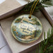 A decorative ornament in a box, featuring a lighthouse and a scenic landscape with a body of water.