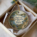 A decorative ornament in a white box, featuring a scenic landscape with a lake, a boat, and a bird.