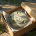 A decorative ornament in a box, featuring a scenic landscape with a lighthouse, trees, and a body of water.