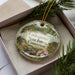 A decorative ornament in a box, featuring a scenic landscape with a river, trees, and wildlife.