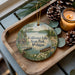 A decorative ornament featuring a serene lakeside scene with a cabin, pine cones, and a candle on a wooden tray.