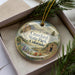 A decorative ornament in a box, featuring a scenic landscape with mountains, water, and a small building.