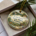 A decorative ornament in a box, featuring a scenic landscape with a river, mountains, and wildlife.