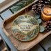 A decorative ornament with a scenic image of Crooked River State Park, Georgia, featuring a waterfall, pine trees, and flowers, is displayed on a wooden tray alongside a lit candle and pine cones.