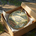 A decorative ornament in a box, featuring a scenic landscape with a river, trees, and a waterfall.