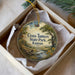 A decorative ornament in a white box, featuring a scenic landscape with a river, trees, and flowers.