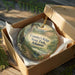 A decorative ornament in a cardboard box, featuring a scenic landscape with a river, mountains, and wildlife.