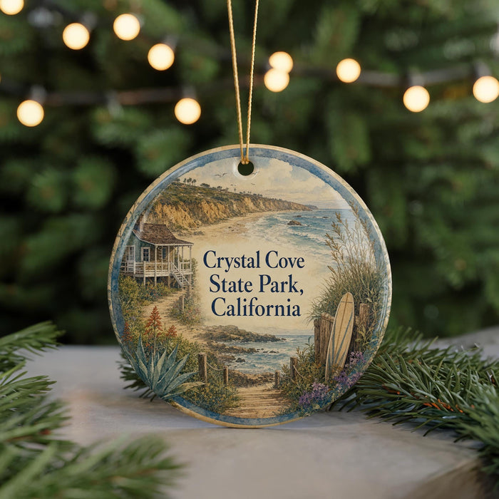 A round ornament featuring a scenic coastal landscape with a house, palm trees, and a surfboard, hanging in front of a blurred background with Christmas lights.
