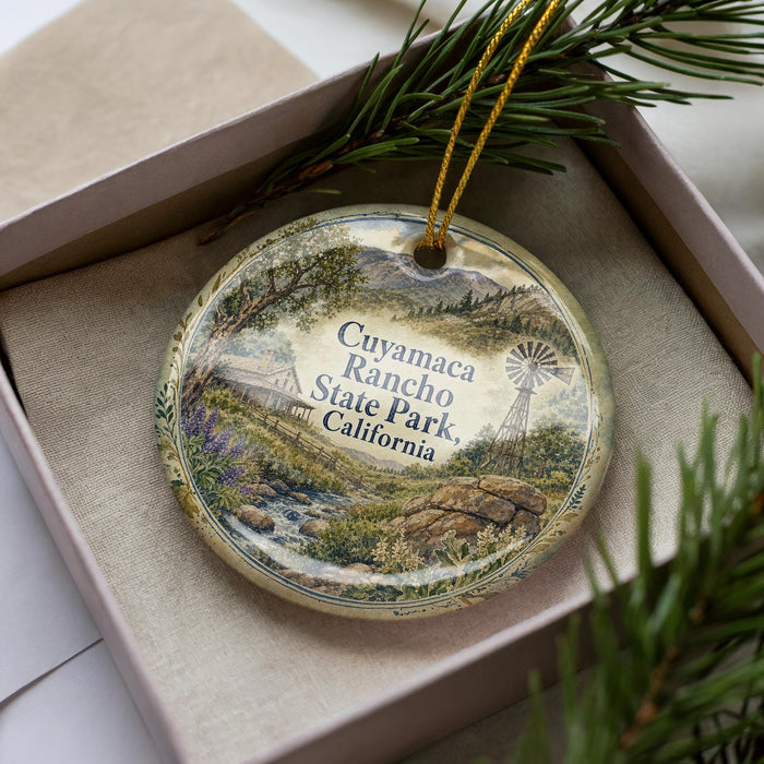 A decorative ornament with a landscape scene featuring a windmill, trees, and a river, placed in a box with pine branches.
