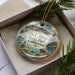 A decorative ornament in a box, featuring a scenic landscape with mountains, water, and pine trees.