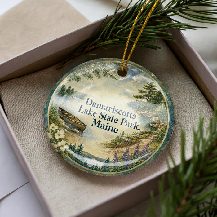 A decorative ornament with a lake and forest scene, featuring a canoe and pine trees, is displayed in a box.