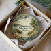 A decorative ornament in a white box, featuring a scenic landscape with a lake, pine trees, and a boat on the water.