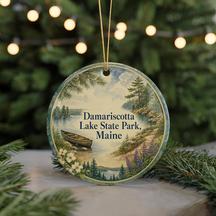 A round ornament with a painting of a lake and pine trees, featuring a boat on the water, hangs in front of a Christmas tree with twinkling lights.