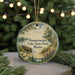 A round ornament with a painting of a lake and pine trees, featuring a boat on the water, hangs in front of a Christmas tree with twinkling lights.