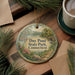 A decorative ornament with a scenic landscape of Day Pond State Park, Connecticut, featuring a lake, trees, and a bridge. The ornament is hanging from a string and is placed on a wooden surface, surrounded by pine branches and a cup of coffee.