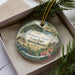 A decorative ornament in a box, featuring a scenic landscape with pine trees, flowers, and a body of water.