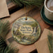 A decorative ornament featuring a scenic image of DeSoto State Park, Alabama, with a waterfall and a bridge, hanging on a wooden surface alongside a cup of coffee and some pine branches.