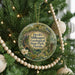 A round ornament hanging on a Christmas tree, featuring a scenic landscape with a winding path and a bird in flight.