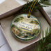 A decorative ornament in a box, featuring a scenic landscape with a river and trees, with the text "Dinosaurs State Park, Connecticut" printed on it.
