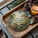A decorative ornament with a scenic image of Dinosaur State Park, Connecticut, featuring a river, trees, and a bridge. The ornament is placed on a wooden tray, with pine cones and a lit candle nearby.