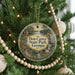 A round ornament hanging on a Christmas tree, featuring a scenic landscape with a house, trees, and a river.